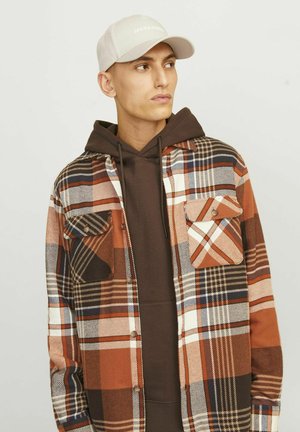Gesp een geraffineerde shirt in oranje, bruin en marineblauw, met twee borstzakken en een knoopsluiting over een bruine hoodie. Het model draagt ook een lichte pet.
