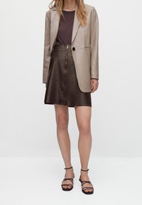 Gonna in pelle marrone con zip frontale, abbinata a un blazer in pelle beige chiaro e a una maglietta marrone scuro. Sandali neri con cinturini completano il look.