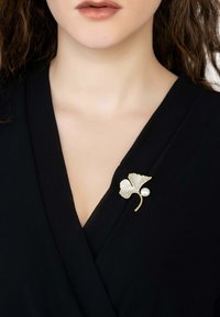 Gold-tone brooch shaped like a ginkgo leaf, adorned with clear crystals and a pearl accent, pinned on a black garment.