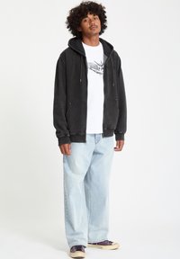 Sudadera negra con cremallera y textura suave, con dos bolsillos frontales, utilizada sobre una camiseta blanca. El modelo lleva unos jeans holgados de color azul claro y zapatillas moradas.