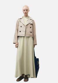 Bežová skrátená bunda s dvojradovým zapínaním a čiernymi gombíkmi cez svetlozelené maxi šaty. Hnedé sandále a modrá taška dopĺňajú outfit.
