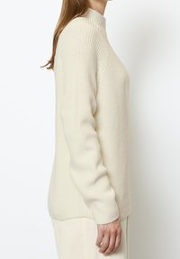 Pull tricoté couleur crème avec un col montant, des manches longues et une texture côtelée. Présente une coupe décontractée et des coutures d'épaule tombantes.