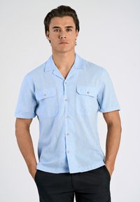 Vaaleansininen lyhythihainen paita, jossa on hienovarainen rakenne, kaksi etutaskua ja napit. Kangas vaikuttaa kevyeltä ja hengittävältä.