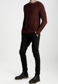 Man with tattooed neck wearing maroon sweater, black slim pants, and black leather boots standing with hands in pockets.