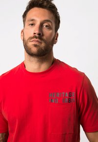 Camiseta de algodón roja con mangas cortas, cuello redondo, bolsillo frontal y gráfico de texto gris que dice "HERITAGE IND. 1880" en el lado izquierdo.