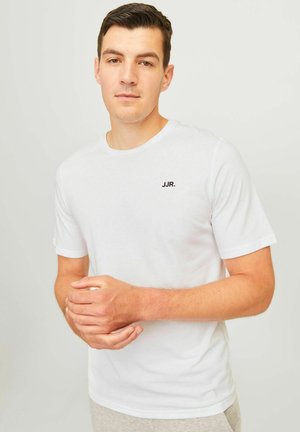 Joven con cabello corto y oscuro, que lleva una camiseta blanca de manga corta con el texto "JJR." en el pecho, de pie frente a un fondo claro.