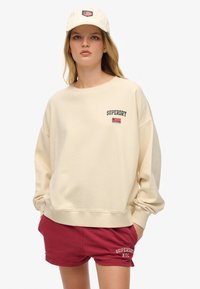 Sudadera beige de gran tamaño con el logo de "SUPERDRY" y un pequeño diseño de bandera; combinada con shorts rojos que presentan un bordado de "SUPERDRY NYC".