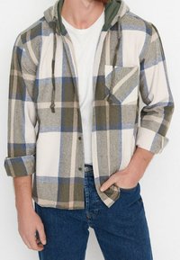 Camicia in flanella a quadri con cappuccio verde, caratterizzata da colori blu, oliva e beige. Chiusura a bottoni, taschino sul petto e maniche arrotolate. Indossata con jeans.