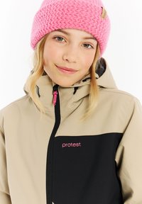 Beige en zwarte jas met roze accenten, rits aan de voorkant, hoge kraag en 'protest'-logo op de linkerkant van de borst. Het model draagt een roze gebreide muts.