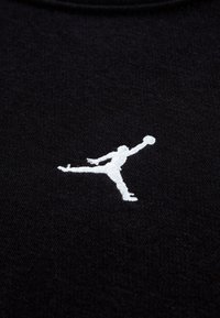 Fekete szövet fehér, hímzett Jumpman logóval, amely egy kosárlabdát tartó ugró alakot ábrázol, tiszta vonalakkal és sima textúrával.