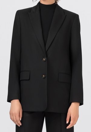 Persoon die een zwarte, op maat gemaakte blazer draagt met twee knopen, klepzakken en een zwart shirt eronder, staand tegen een effen achtergrond.