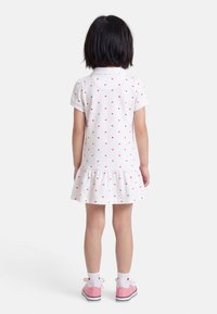 Παιδί που φαίνεται από πίσω, φορώντας ένα λευκό φόρεμα με κόκκινες καρδιές, λευκές κάλτσες με καρδιές και ροζ αθλητικά παπούτσια σε λευκό φόντο.