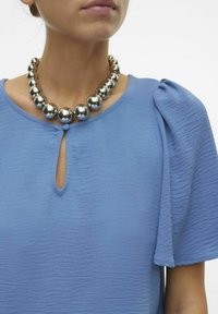 Blusa azzurro chiaro in tessuto texturizzato con scollatura a chiave e maniche a sbuffo, accentuata da una collana di perle metalliche chunky.