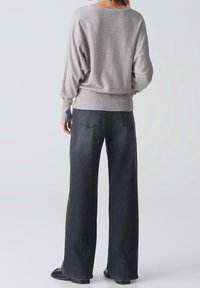 Kobieta ubrana w jasnoszary sweter z długim rękawem, ciemne dżinsy o szerokich nogawkach oraz czarne buty, stojąca tyłem na jednolitym tle.