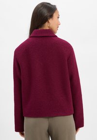 Burgundy wool blend jacket with a collar, dropped shoulders, and a textured finish. Features a relaxed fit, ending at the waist.