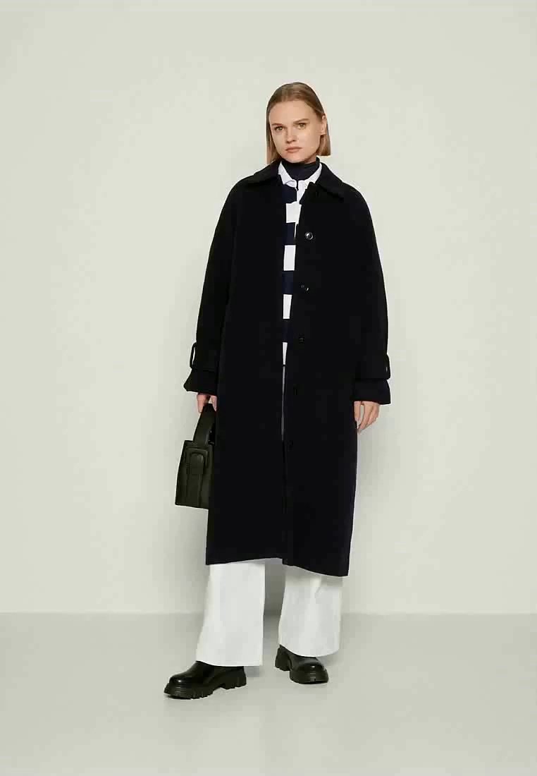 Femme portant un long manteau noir sur un haut rayé noir et blanc, un pantalon blanc, des chaussures noires, tenant un sac à main noir, se tenant contre un fond uni.