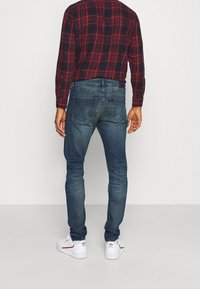 Jeans skinny blu scuro in denim, con leggeri effetti di scolorimento, dettagli di cucitura sulle tasche posteriori, indossati con una camicia a quadri rossa e blu navy.