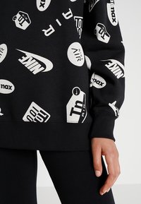 Svart sweatshirt med olika vita logotyper och text, med ribbad nederkant och ärmslut, tillverkad av mjukt bomullsmaterial.