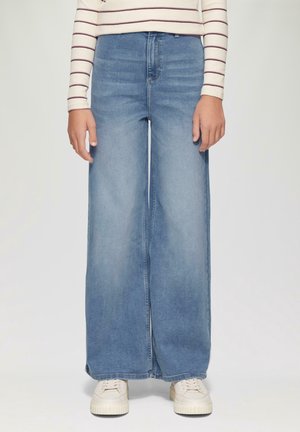 Jeans azzurri a gamba larga con vita alta e un leggero sbiadimento, abbinati a sneakers crema con lacci testurizzati e suola spessa.