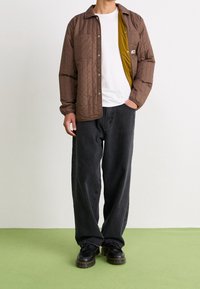 Osoba w brązowej pikowanej kurtce, pod którą ma białą koszulkę, luźnych czarnych dżinsach i czarnych sznurowanych butach, stoi na zielonej podłodze.