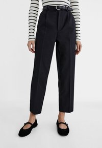 Pantalon tailleur noir à taille haute, avec des plis et une ceinture. Porté avec un haut à manches longues rayé noir et blanc et des ballerines noires.