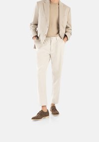 Blazer beige sobre una camisa marrón, combinado con pantalones beige claro y zapatos de ante marrones, con cordones y una suela clara.