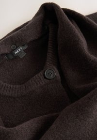Cardigan en laine marron foncé avec des boutons noirs et une étiquette visible portant la mention "NEXT" sur un fond clair.