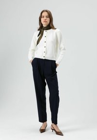 Cardigan en maille blanche avec boutons noirs, associé à un pantalon plissé bleu marine et des chaussures à talons marron. Écharpe noire à motifs autour du cou.