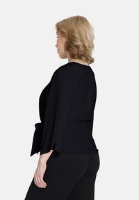 Top nero avvolgente con maniche a campana, texture morbida e un nodo in vita. Visto di lato, abbinato a pantaloni neri.