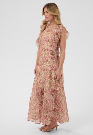 Femme aux longs cheveux blonds portant une robe maxi rose fleurie sans manches avec détails à volants et des sandales à talons beiges, debout de côté.