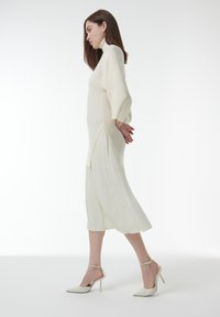 Lange, geplooide, witte jurk met een hoge hals en geribbelde textuur, gecombineerd met witte hoge hakken. De mouwen zijn wijd en vloeiend.