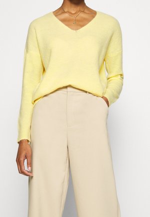 Persona que lleva un suéter amarillo suave con cuello en V metido en unos pantalones anchos de talle alto de color beige, con una mano en el bolsillo.
