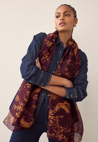 Écharpe bordeaux avec un motif floral orange, en tissu léger, drapée sur une chemise en jean bleue. Finition texturée visible.