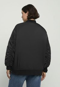 Chaqueta bomber negra con mangas fruncidas, puños y dobladillo de canalé. Material suave y brillante. Vista trasera que muestra el cuello y un ajuste relajado.