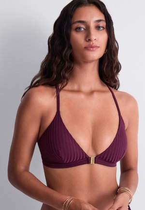 Femme aux longs cheveux ondulés foncés, portant un haut de bikini côtelé violet profond avec une fermeture dorée, posant devant un fond gris uni.