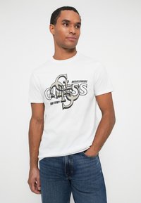 Mand iført hvid kortærmet t-shirt med overlappende "GUESS" logo grafik og blå jeans, stående med den ene hånd i lommen.