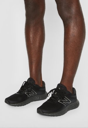 Paar schwarze New Balance Laufschuhe an dunkelhäutigen Beinen, die auf einem schlichten hellgrauen Hintergrund stehen.