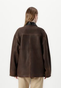 Brūna ādas jaka ar melnu apkakli, pārlieku liela izmēra, redzamām šuvēm un gludu tekstūru, skatoties no aizmugures.