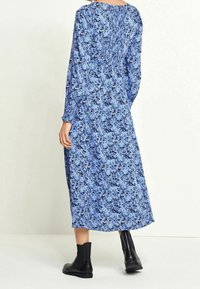 Femme portant une robe midi à manches longues bleue à fleurs, associée à des bottines noires, debout devant un fond uni clair.