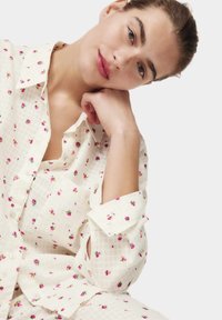 Camicia bianca con bottoni, caratterizzata da un motivo a griglia e piccoli dettagli floreali rosa. Vestibilità rilassata con maniche arrotolate e taschino applicato.