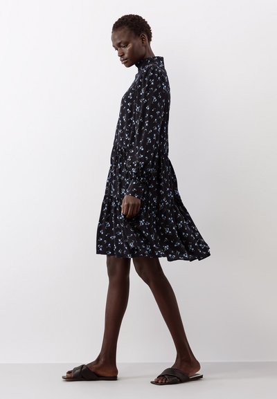 Vestido preto floral com mangas longas, gola alta e saia em camadas. Apresenta pequenas flores azul claro no tecido. Usado com chinelos pretos.