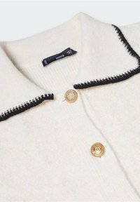 Bílý kardigan z vlněné směsi s černým prošitým límcem, s předním zapínáním na zlatý knoflík a připevněnou štítkem s péčí.