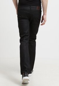 Vaqueros de mezclilla negra con corte recto, que presentan bolsillos traseros y un parche de cuero marrón. Llevados con zapatillas negras sobre un fondo claro.