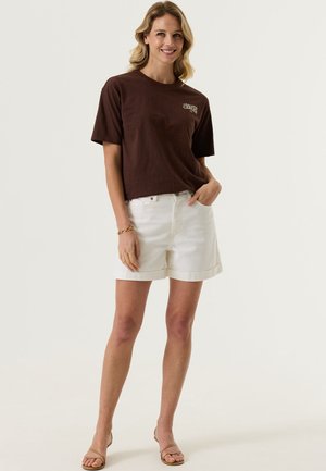 Femme aux cheveux blonds ondulés portant un t-shirt marron à manches courtes avec un logo, un short blanc taille haute et des sandales beiges, debout et souriante.