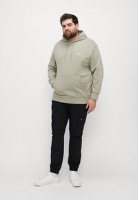 Jasnokhaki hoodie z przednią kieszenią, białym logo i sznurkami, zestawiony z czarnymi spodniami cargo i białymi trampkami. Gładka tekstura.