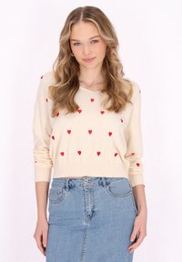 Maglione beige con motivi a cuore rossi, caratterizzato da un collo a V e maniche lunghe. Indossato con una gonna di jeans blu chiaro.