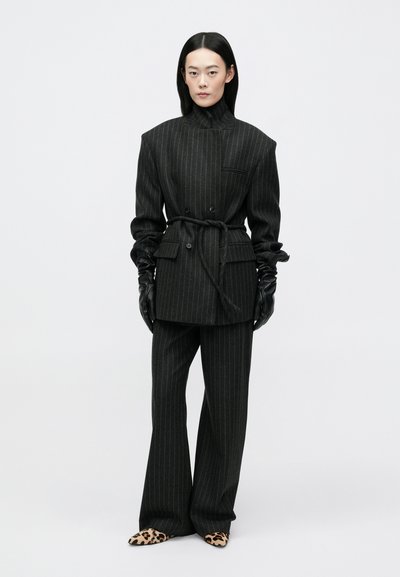 Fekete csíkos zakó magas nyakkal, övvel és dupla gombokkal, amelyet hozzáillő nadrággal és állati mintás magassarkú cipőkkel párosítanak.