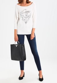 Weißes, schulterfreies Oberteil mit schwarzem Schriftzug und floralem Design, kombiniert mit dunkelblauen Skinny-Jeans und schwarzen High Heels, dazu eine schwarze Handtasche haltend.