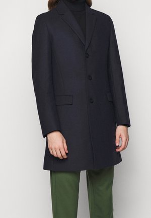 Personne portant un manteau noir à simple boutonnage sur un col roulé noir avec un pantalon vert, debout devant un fond clair uni.