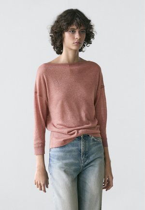 Jeune personne aux cheveux bouclés portant un haut rose à manches longues et amples, ainsi qu'un jean bleu délavé, se tenant devant un fond clair uni.